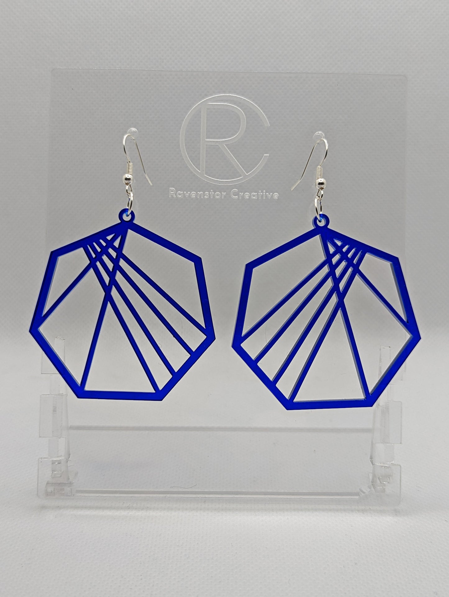 Shows a pair of earrings that have silver ear wire hook fixings. They are Geometric Heptagons with lines crisscrossing through the middle. They are made from Translucent Blue Acrylic and are against a white background on see through stand.