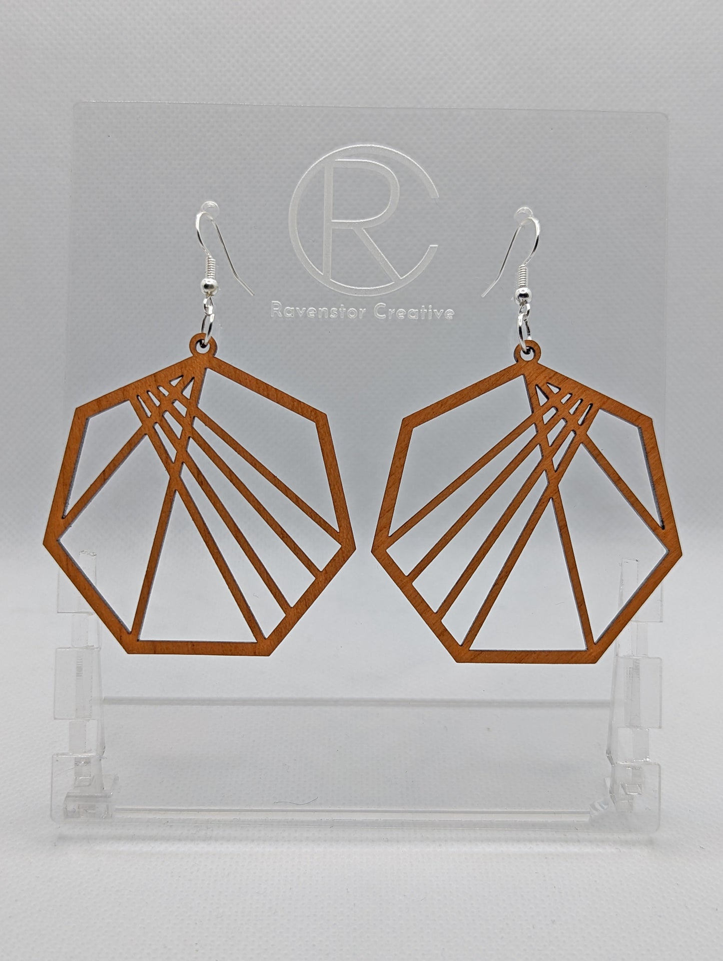 Shows a pair of earrings that have silver ear wire hook fixings. They are Geometric Heptagons with lines crisscrossing through the middle. They are made from Cherry Wood and are against a white background on see through stand.