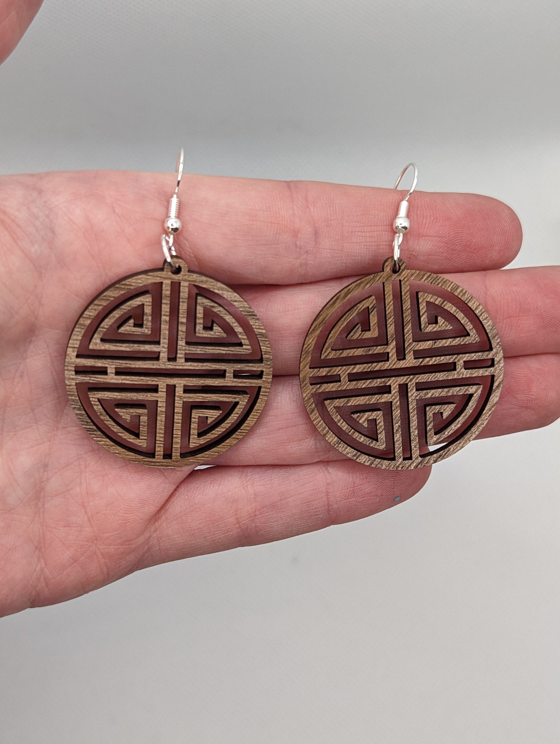 Shows a pair of Geometric Korean Inspired Circle earrings with an intricate pattern like a maze on a hand showing the size against a white background, they are two finger widths in length and two wide. They are Walnut wood and have silver fixings.