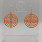 Shows a pair of acrylic earrings that have rose gold ear wire hook fixings. They are Geometric Korean Inspired Circle earrings with an intricate pattern like a maze. They are orange colour and are against a white background on see through stand.