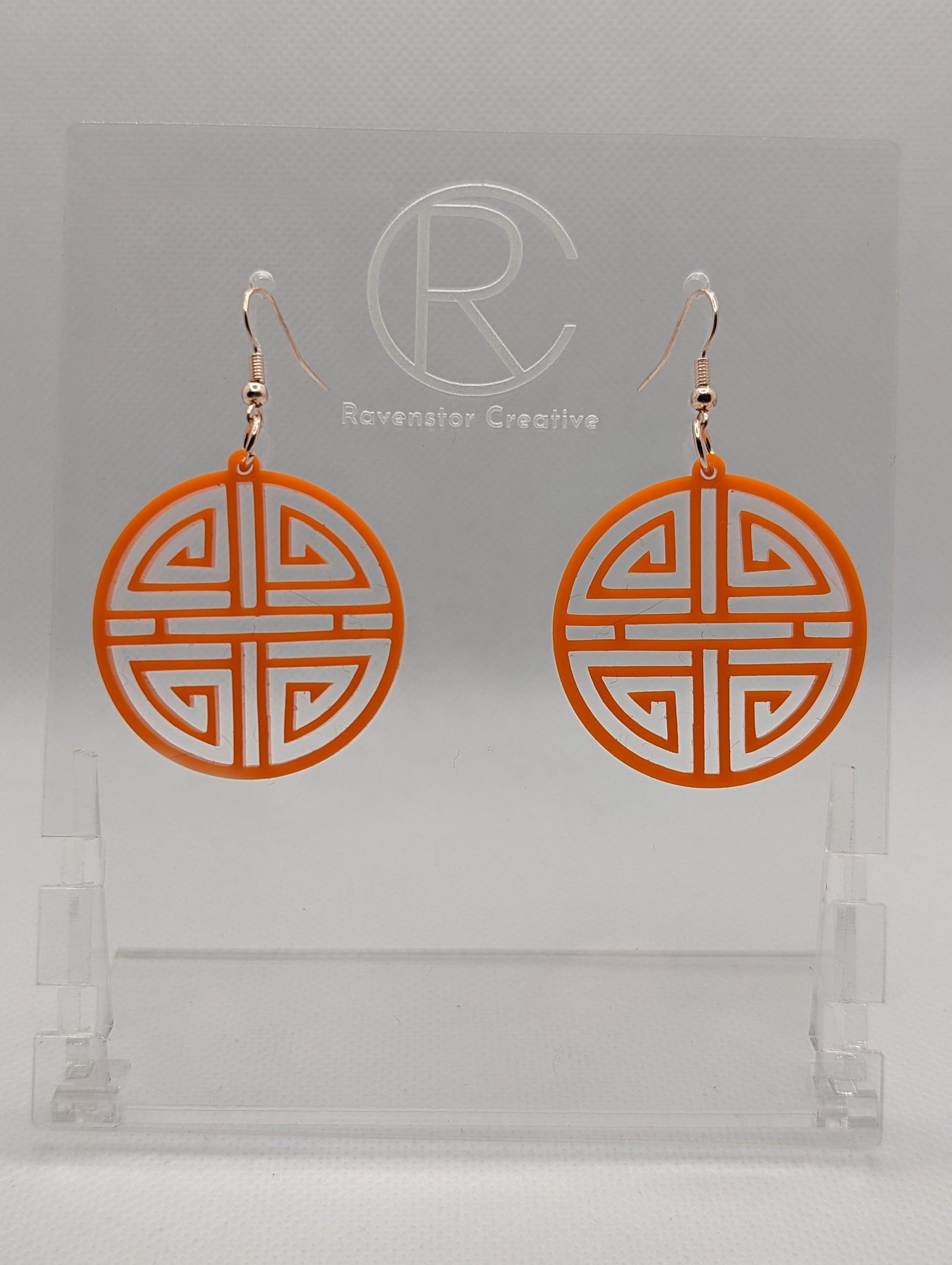 Shows a pair of acrylic earrings that have rose gold ear wire hook fixings. They are Geometric Korean Inspired Circle earrings with an intricate pattern like a maze. They are orange colour and are against a white background on see through stand.