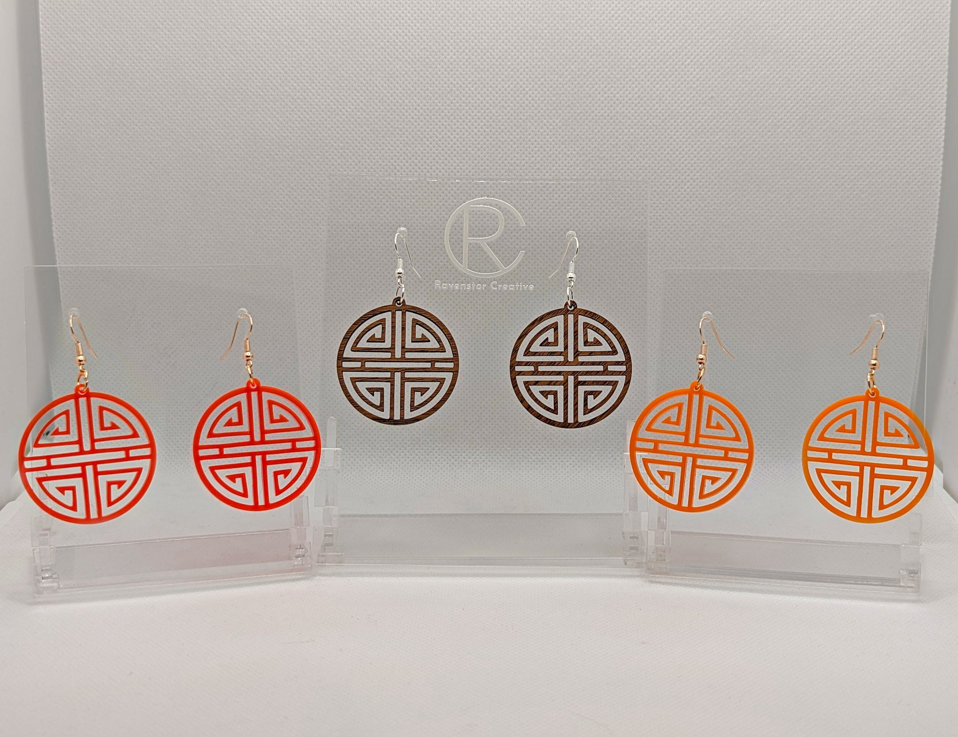 Shows three pairs of earrings that have ear wire hook fixings. They are Geometric Korean Inspired Circle earrings with an intricate pattern. The colours are neon pink, walnut wood and orange. They are against a white background on see through stands.