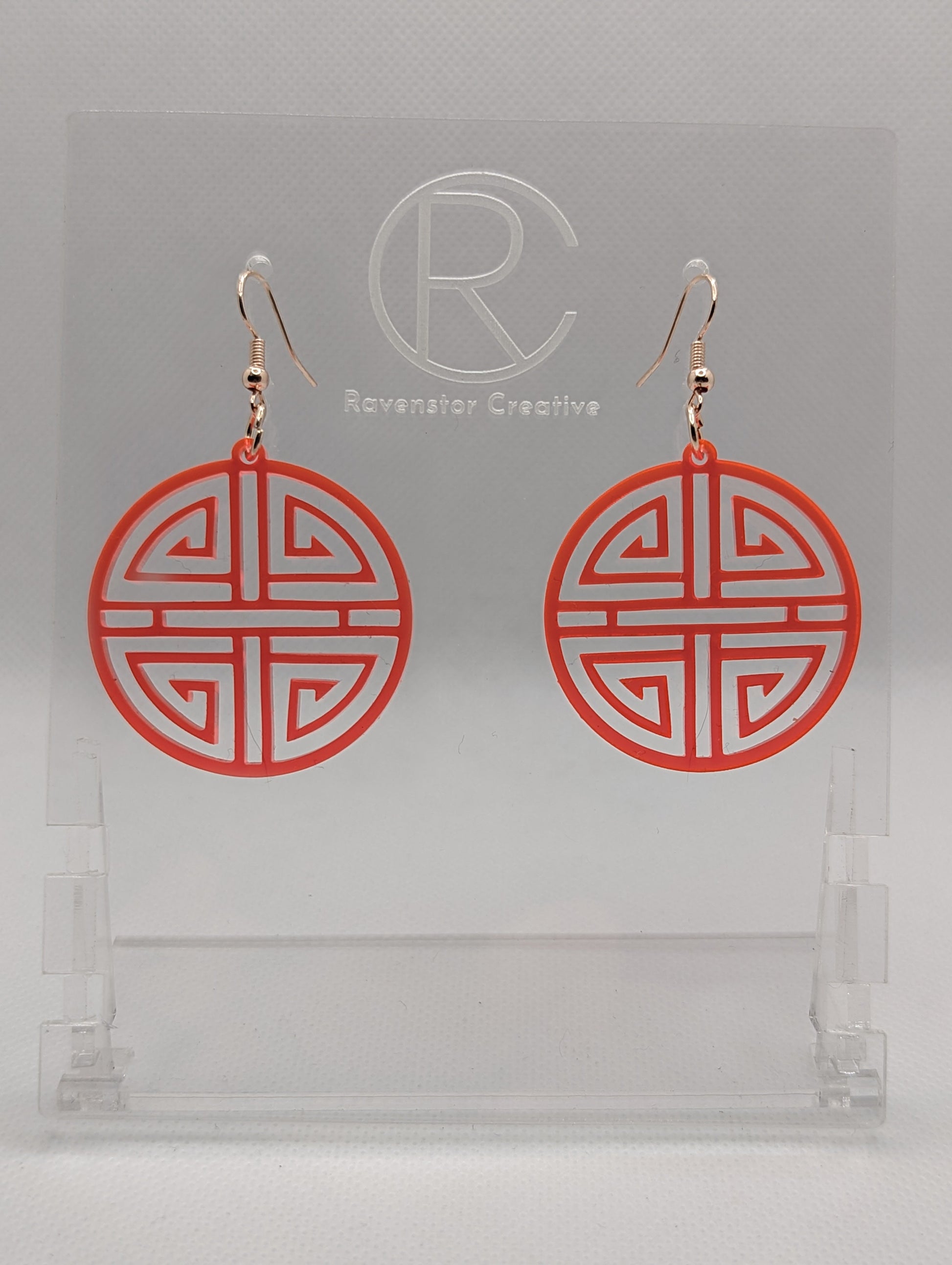 Shows a pair of acrylic earrings that have rose gold ear wire hook fixings. They are Geometric Korean Inspired Circle earrings with an intricate pattern like a maze. They are neon pink colour and are against a white background on see through stand.