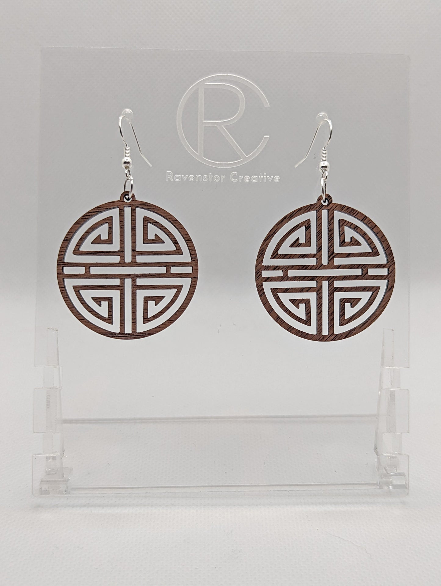 Shows a pair of earrings that have silver ear wire hook fixings. They are Geometric Korean Inspired Circle earrings with an intricate pattern like a maze. They are walnut wood and are against a white background on see through stand.