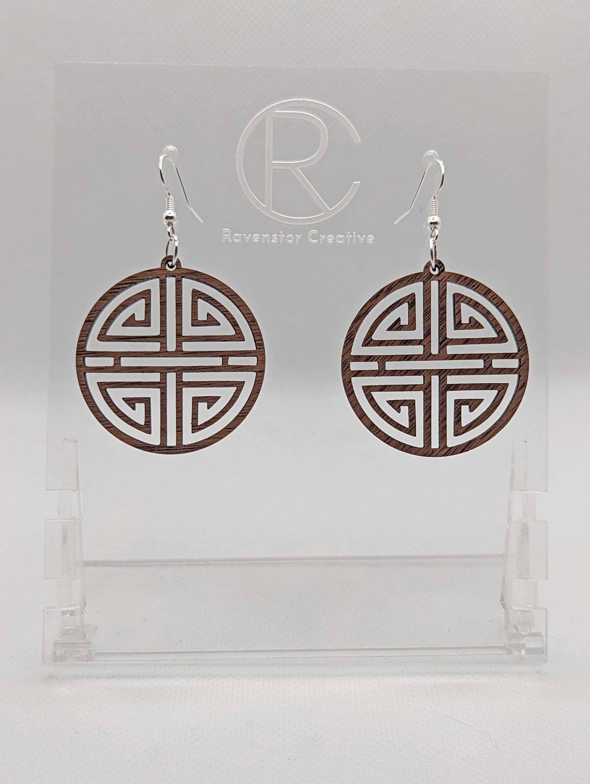 Shows a pair of earrings that have silver ear wire hook fixings. They are Geometric Korean Inspired Circle earrings with an intricate pattern like a maze. They are walnut wood and are against a white background on see through stand.