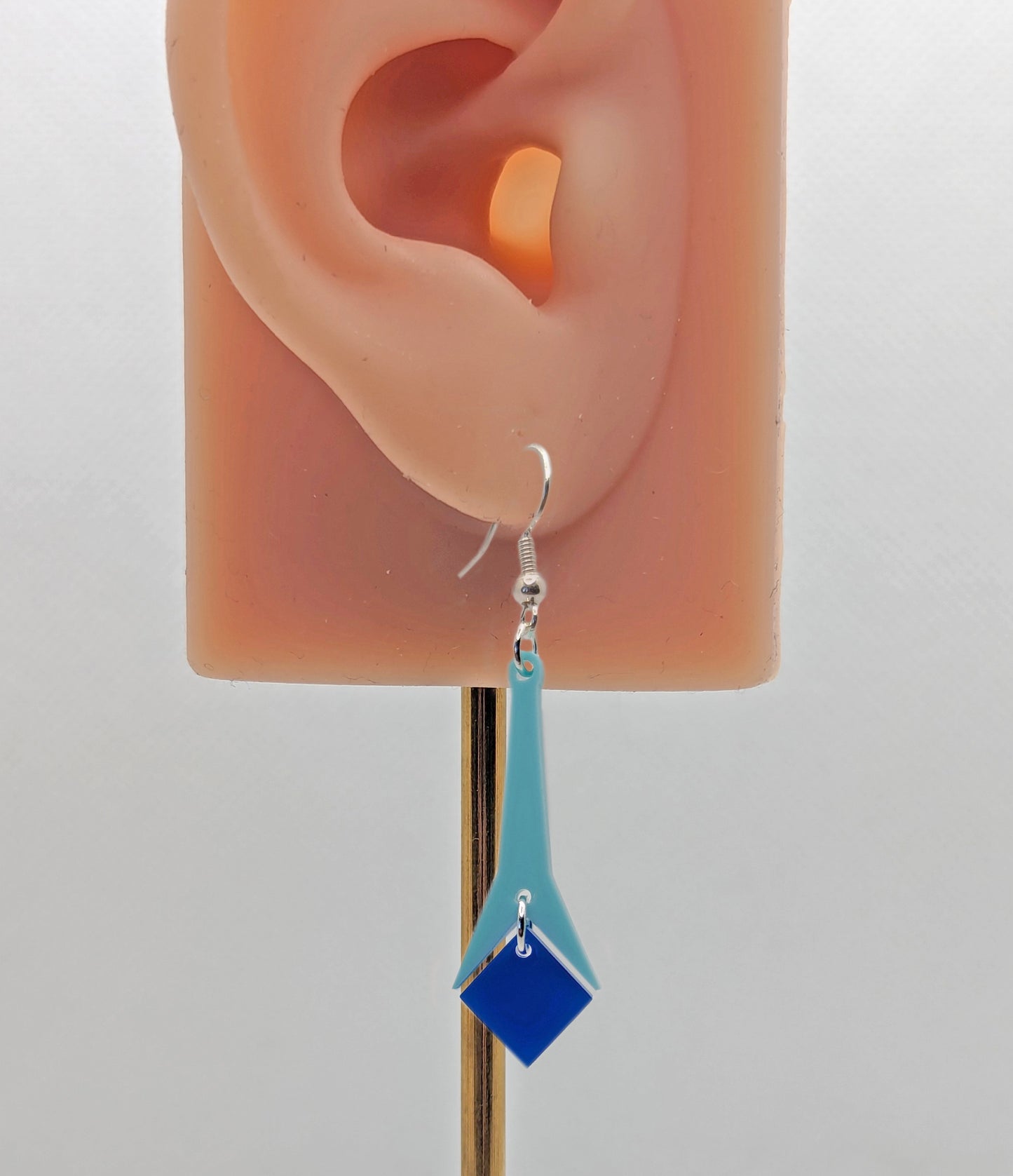 Shows an earring with silver ear wire hook fixings. It is a slim geometric teardrop shape in two parts on an ear showing thier size. It is made from spearmint and blue acrylic.