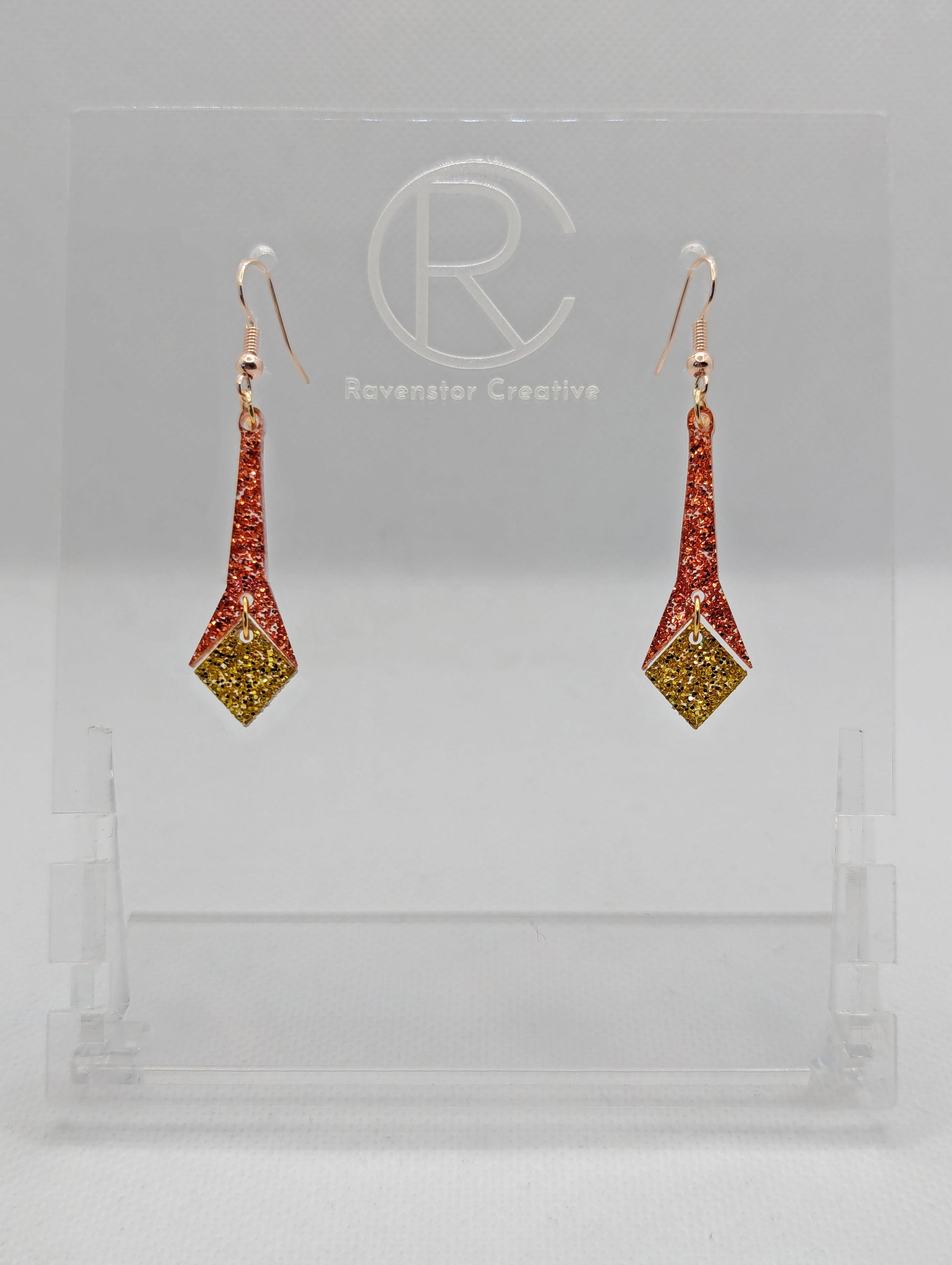 Shows a pair of earrings with rose gold ear wire hook fixings. They are slim teardrop geometric shapes in two parts with a diamond shape dangling under. The colours are Copper Glitter top and gold glitter diamond. They are on a clear stand