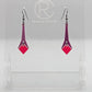 Shows a pair of earrings with black ear wire hook fixings. They are slim teardrop geometric shapes in two parts with a diamond shape dangling under. The colours are grape top and hot pink diamond. They are on a clear stand against a white backing