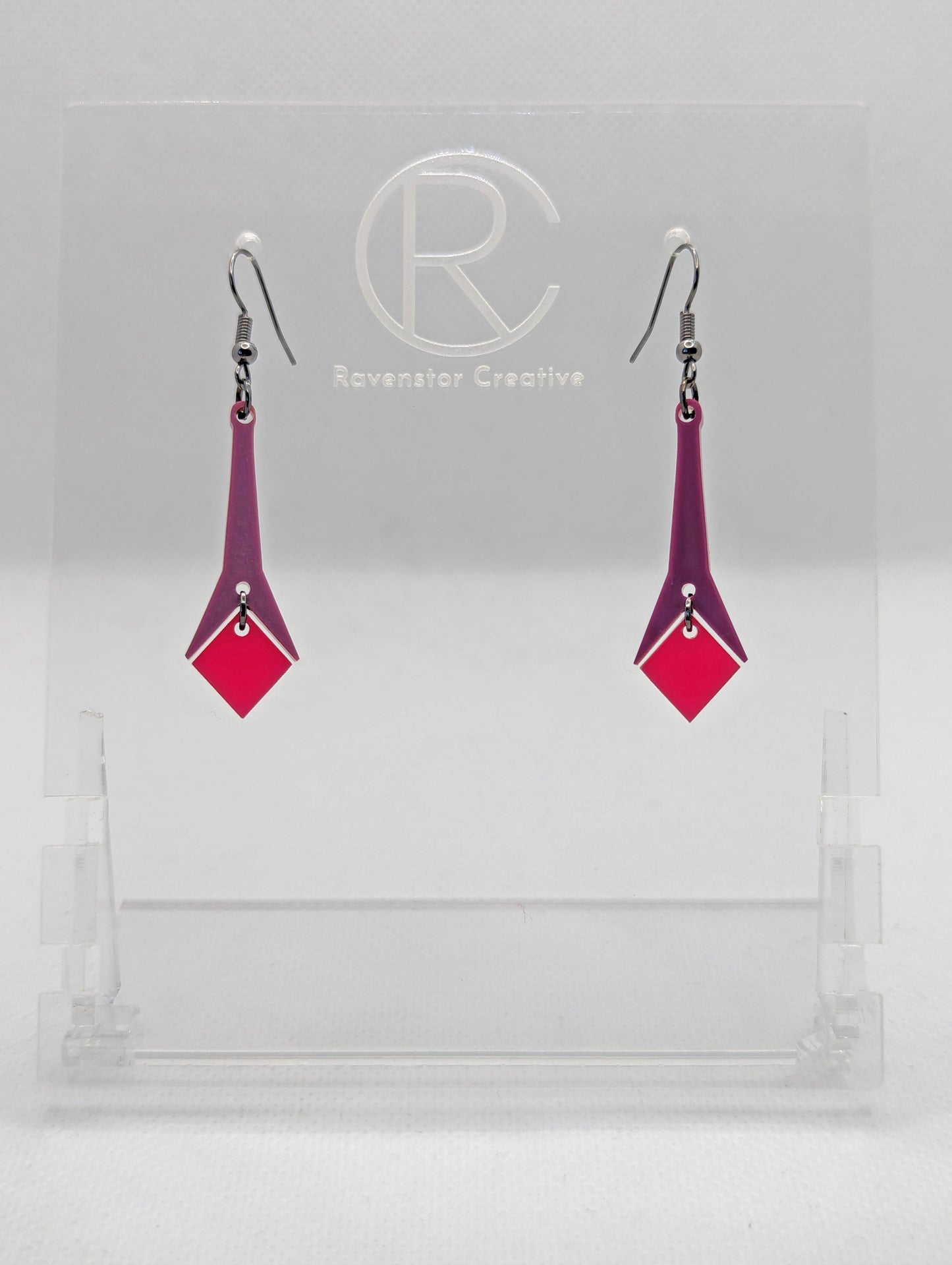 Shows a pair of earrings with black ear wire hook fixings. They are slim teardrop geometric shapes in two parts with a diamond shape dangling under. The colours are grape top and hot pink diamond. They are on a clear stand against a white backing