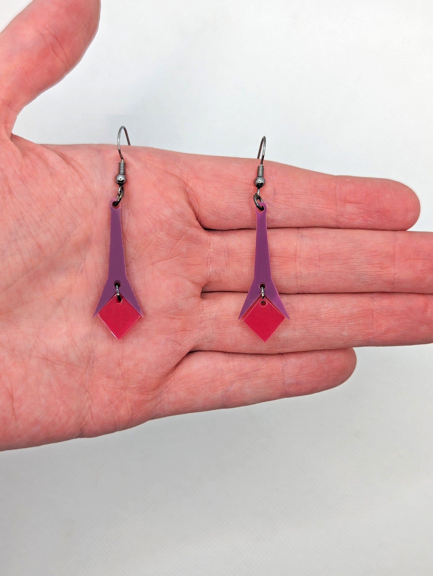 Shows a pair of earrings with ear wire hook fixings. They are slim geometric teardrop shapes in two parts on a hand showing thier size, they are two and a half finger widths in length and one wide. The colours are grape top with hot pink bottom.