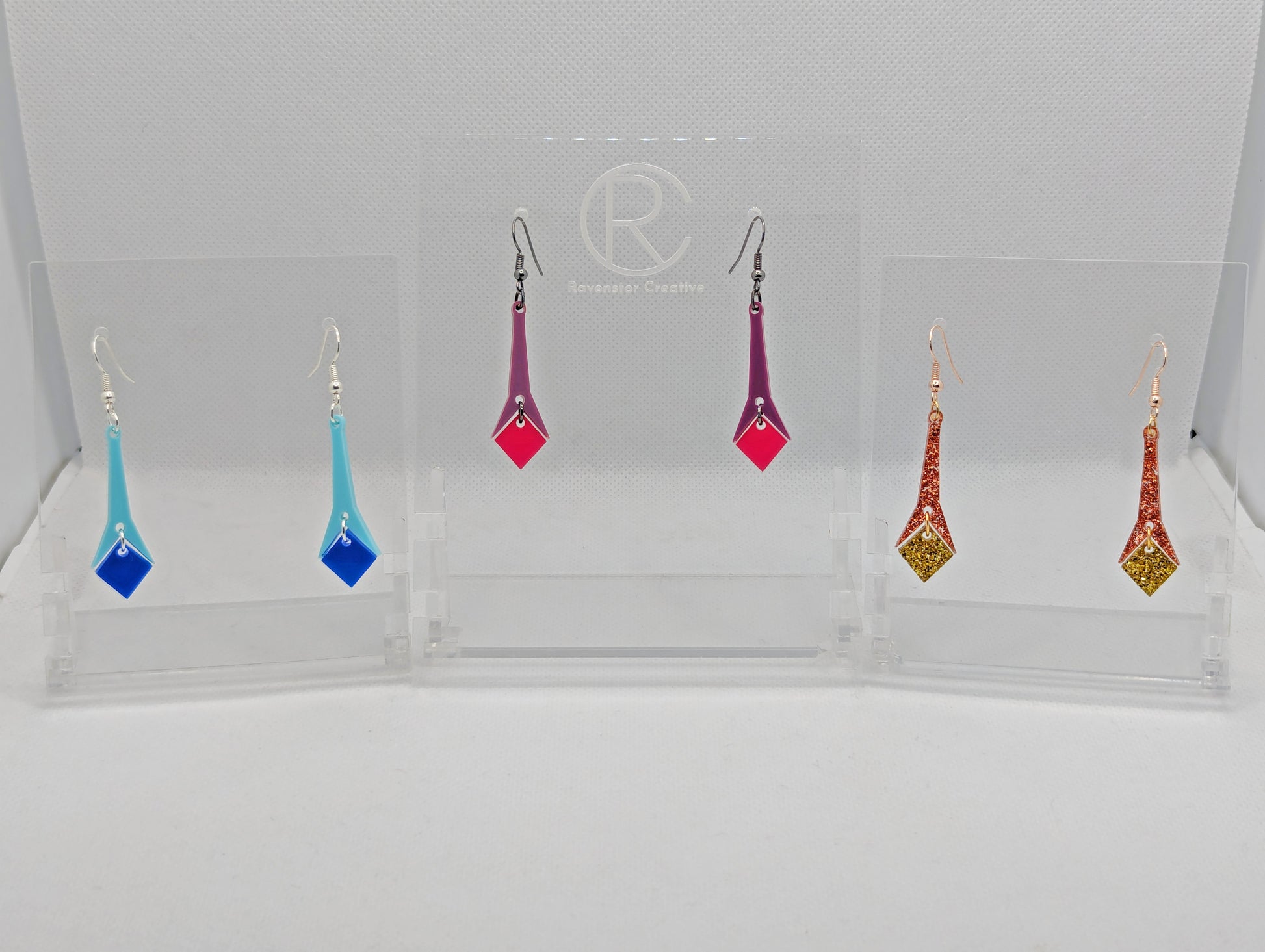 Shows three pairs of 2 part earrings with ear wire hook fixings. They are slim geometric teardrop shapes. The colours are spearmint top and blue bottom, grape top and hot pink bottom, copper glitter top and gold glitter bottom.