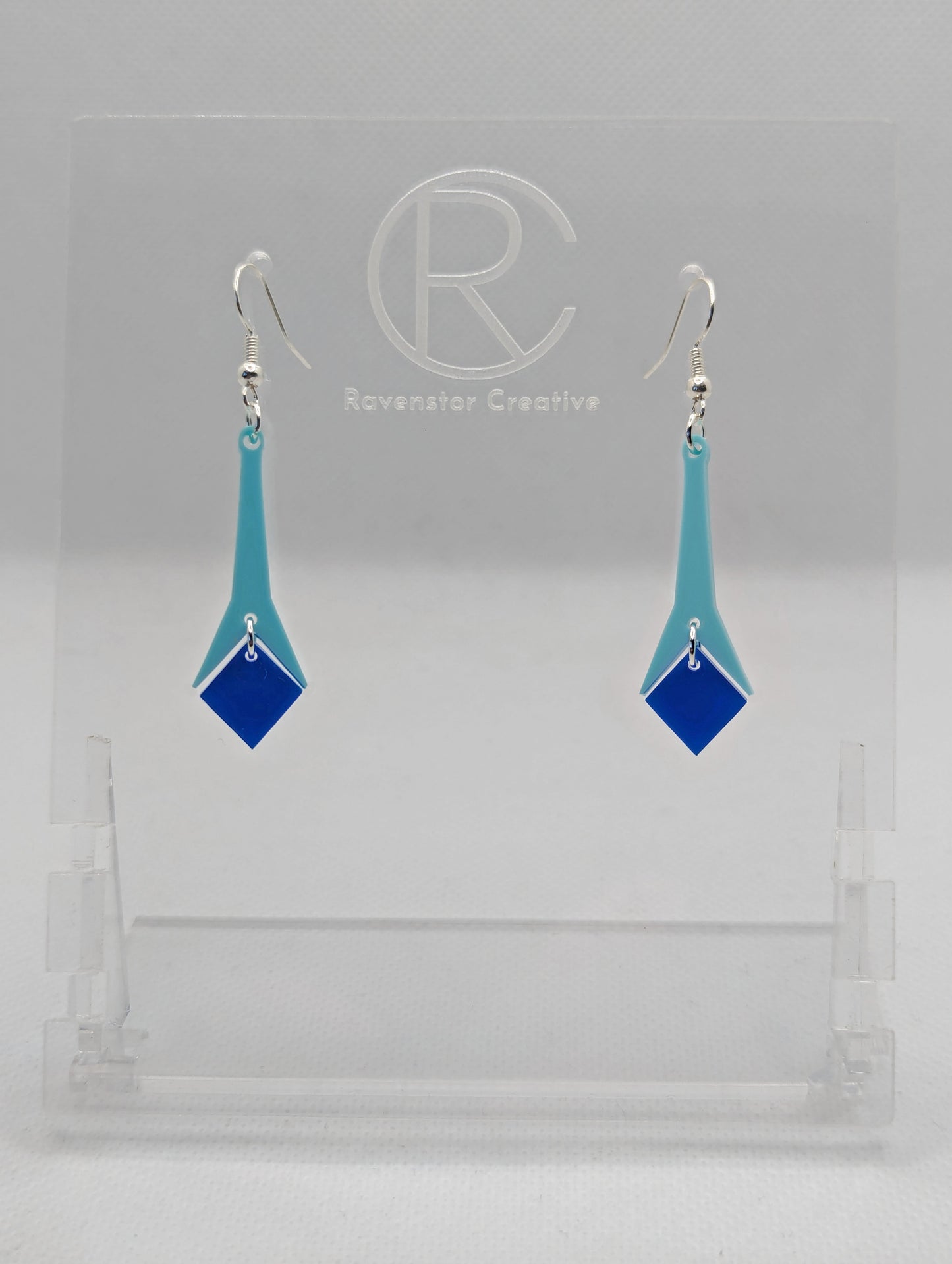 Shows a pair of earrings with silver ear wire hook fixings. They are slim teardrop geometric shapes in two parts with a diamond shape dangling under. The colours are spearmint top and blue diamond. They are on a clear stand against a white backing