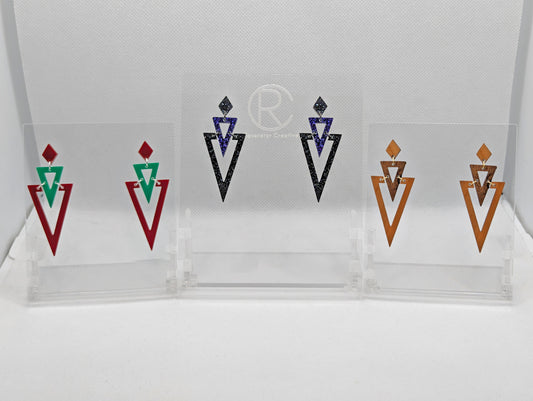 Shows three pairs of geometric themed earrings with stud fixings. They are a diamond shape, with an upside-down triangle under with another triangle under that. The colours are Green, Maroon, Cherry and Walnut wood, Purple and Black / Silver Glitter.