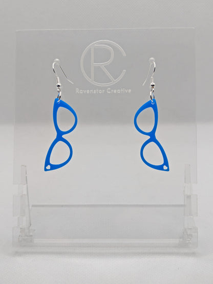 Shows a pair of acrylic earrings that have ear wire hook fixings. They are fab sassy glasses with a heart earrings. They are blue in colour and are against a white background on see through stand with the Ravenstor Creative logo on it.