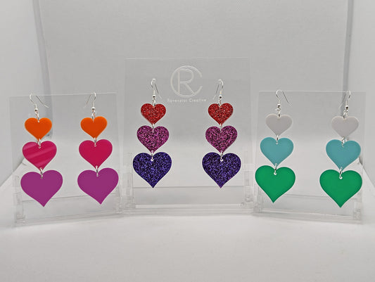 Three pairs of earrings, each has three hearts dangling from each other, the top is the smallest and they get bigger as they go down. Colours are (from the top) orange, Hot pink and grape. Glitters Red, Pink and Purple. White, spearmint and green.