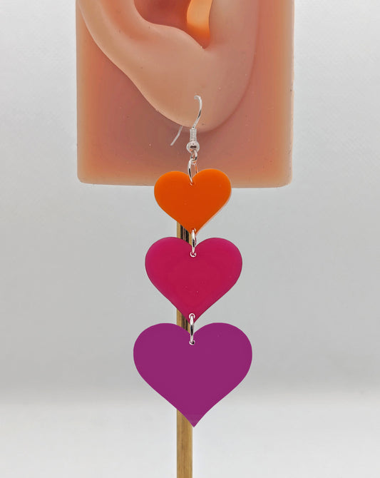 An earrings which has three hearts dangling from each other, the top is the smallest and they get bigger as they go down. It is on an ear to show the size. It is made from Orange, Hot pink and grape acrylic and have silver ear wire hook fixings.