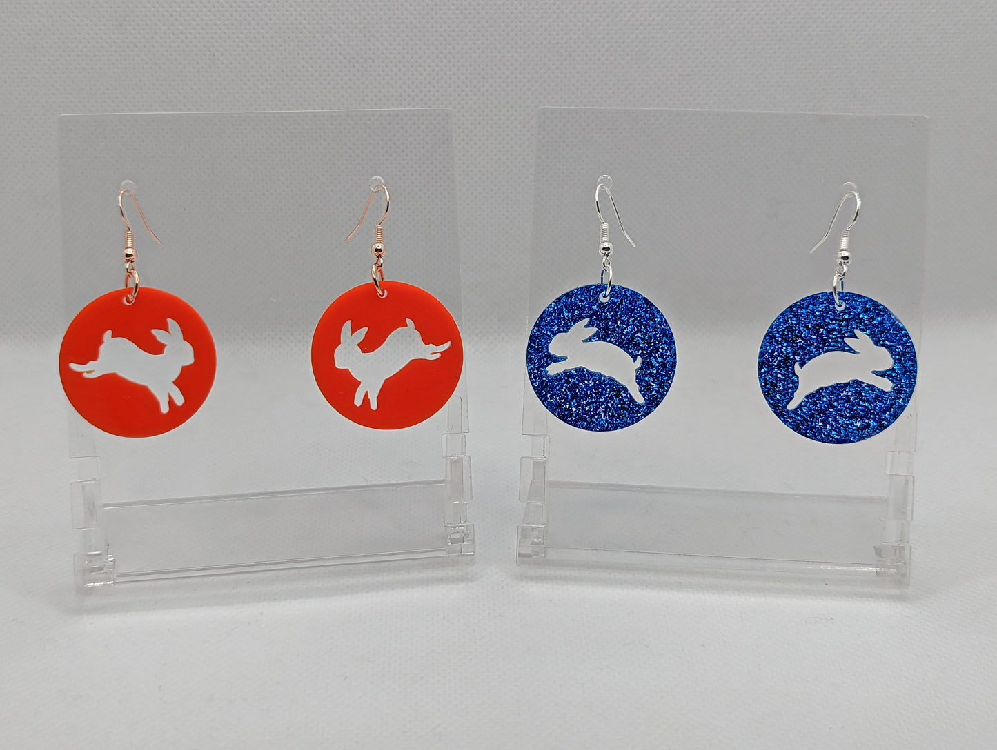 Two pairs of acrylic earrings that have ear wire hook fixings. They are Circles with an hopping Easter bunny / Rabbit cut out each one. The colours are Red and Royal Blue Glitter. They are against a white background on see through stands.