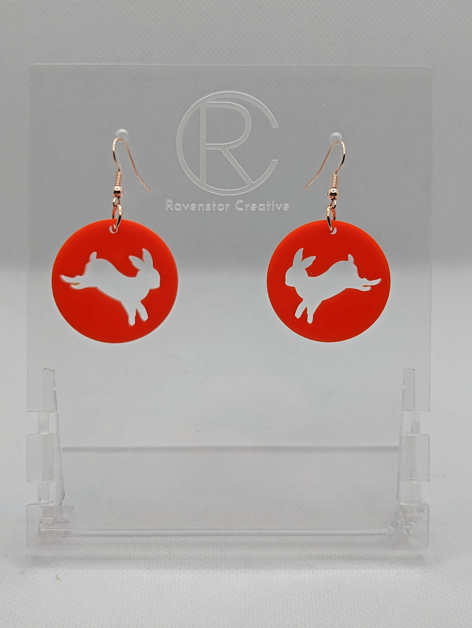 A pair of acrylic earrings that have Rose Gold ear wire hook fixings. They are Circles with an Easter bunny / Rabbit hopping down cut out each one. It is Red colour. They are against a white background on see through stands.