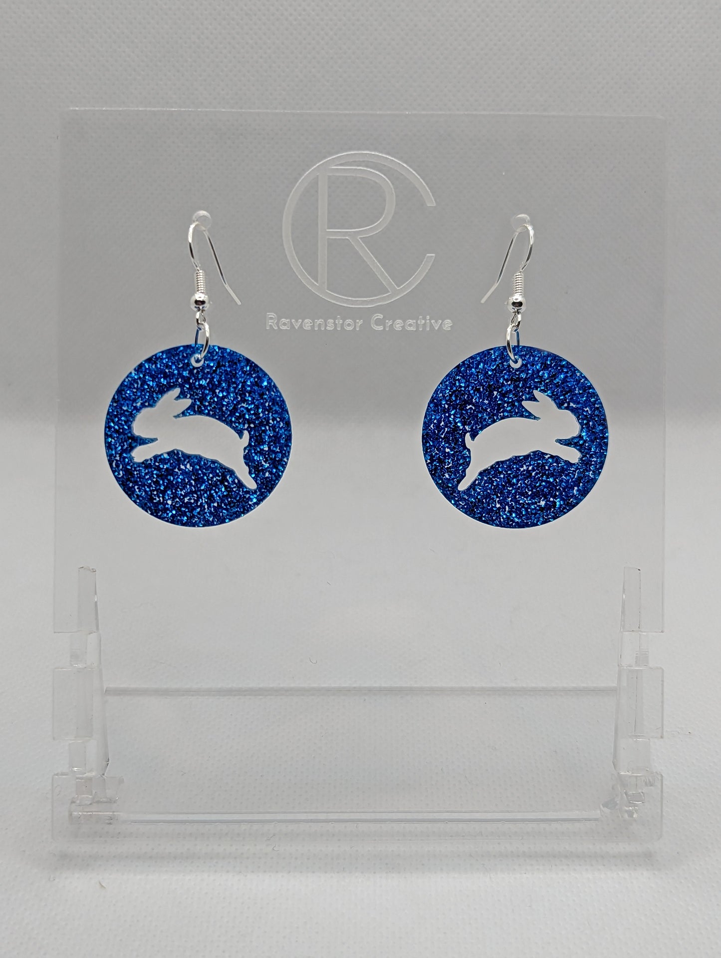 A pair of acrylic earrings that have Silver ear wire hook fixings. They are Circles with an Easter bunny / rabbit hopping up cut out each one. It is Royal Blue Glitter colour. They are against a white background on see through stands.