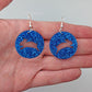 A pair of acrylic earrings that have Silver ear wire hook fixings against a hand to show the size of two finger widths long and same again wide. They are Circles with an Easter bunny / Rabbit hopping cut out each one. It is Royal Blue Glitter colour.