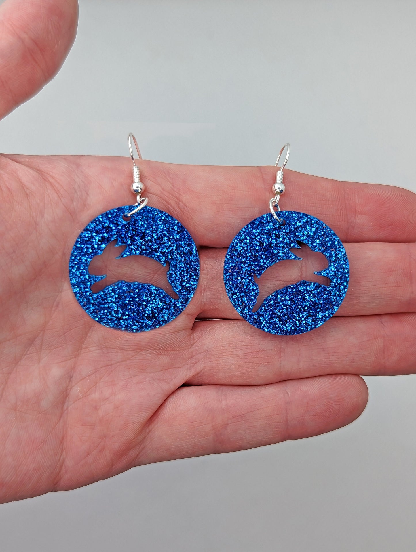 A pair of acrylic earrings that have Silver ear wire hook fixings against a hand to show the size of two finger widths long and same again wide. They are Circles with an Easter bunny / Rabbit hopping cut out each one. It is Royal Blue Glitter colour.