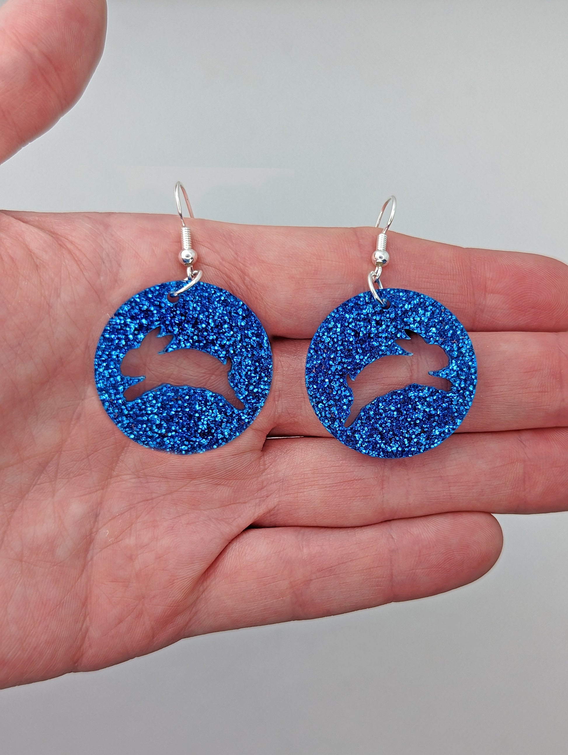 A pair of acrylic earrings that have Silver ear wire hook fixings against a hand to show the size of two finger widths long and same again wide. They are Circles with an Easter bunny / Rabbit hopping cut out each one. It is Royal Blue Glitter colour.