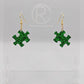 Shows a pair of acrylic earrings with gold ear wire hook fixings. They are Jigsaw Game Piece Shaped. The colour is Green Glitter. They are against a white background on a clear stand with the Ravenstor Creative logo on it.