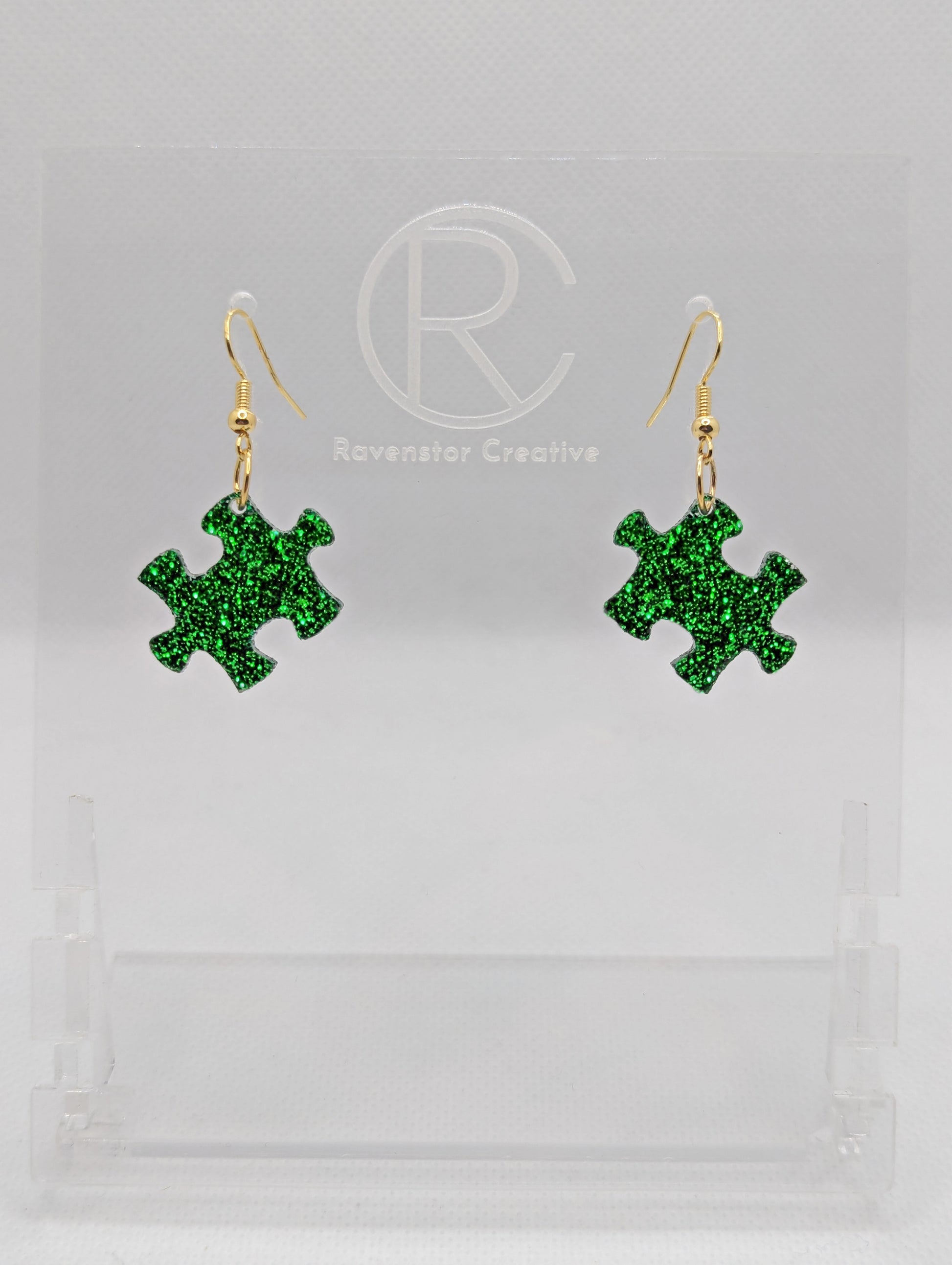 Shows a pair of acrylic earrings with gold ear wire hook fixings. They are Jigsaw Game Piece Shaped. The colour is Green Glitter. They are against a white background on a clear stand with the Ravenstor Creative logo on it.