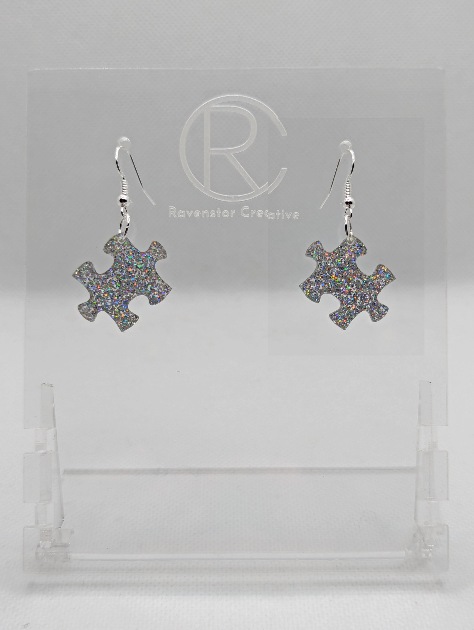 Shows a pair of acrylic earrings with silver ear wire hook fixings. They are Jigsaw Game Piece Shaped. The colour is Holographic Glitter. They are against a white background on a clear stand with the Ravenstor Creative logo on it.