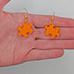 Shows a pair of acrylic earrings with silver ear wire hook fixings. They are Jigsaw Game Piece Shaped. The colour is Orange. They are against a hand showing the size of one finger width in length and slightly larger width.