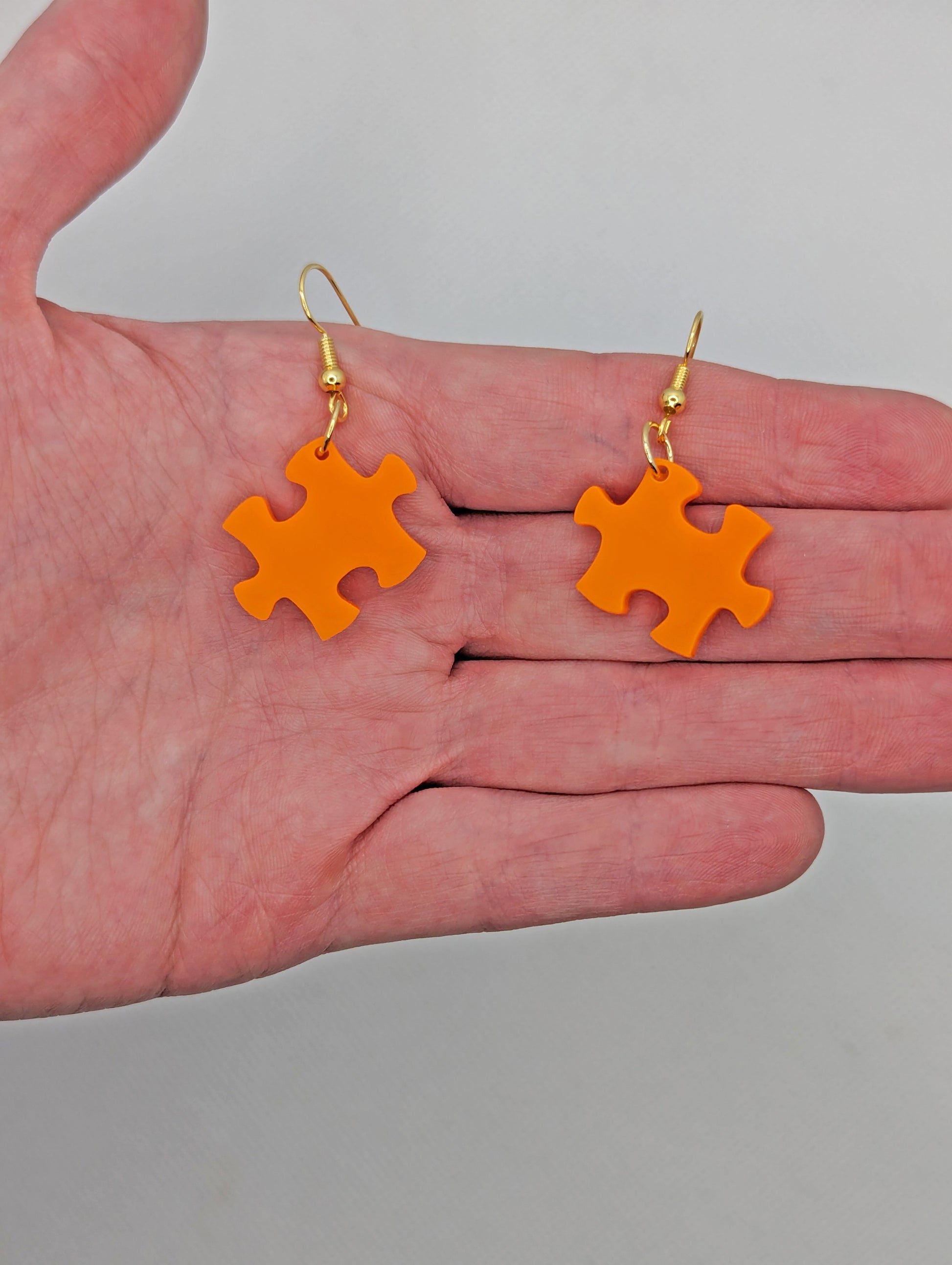 Shows a pair of acrylic earrings with silver ear wire hook fixings. They are Jigsaw Game Piece Shaped. The colour is Orange. They are against a hand showing the size of one finger width in length and slightly larger width.