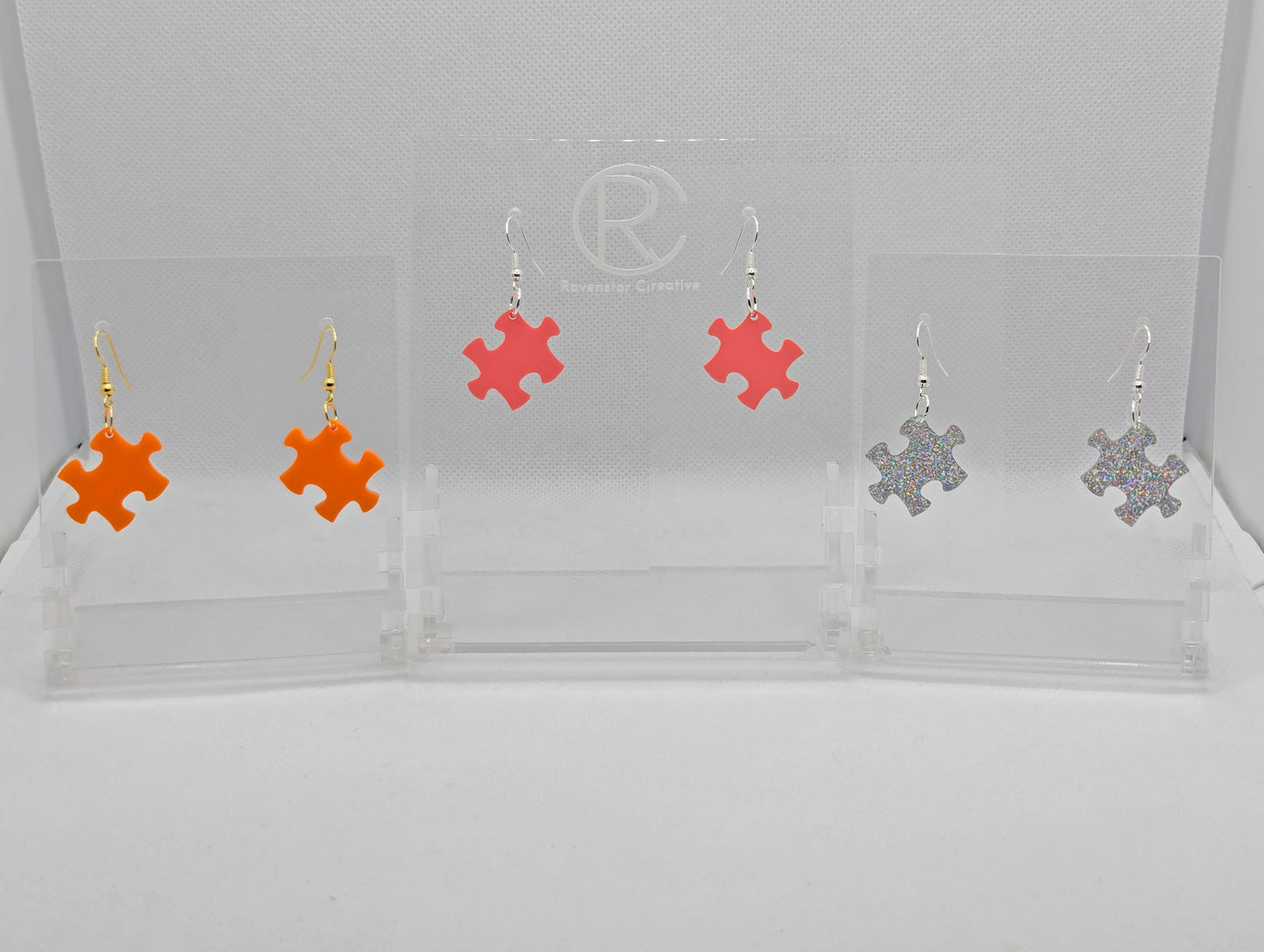 Shows three pairs of acrylic earrings with ear wire hook fixings. They are Jigsaw Game Piece Shaped. The colours are Orange, Holographic Glitter and Sherbet Pink. They are against a white background on a clear stand.