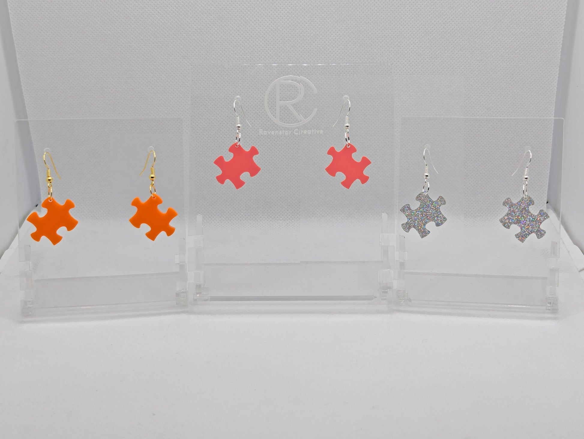 Shows three pairs of acrylic earrings with ear wire hook fixings. They are Jigsaw Game Piece Shaped. The colours are Orange, Holographic Glitter and Sherbet Pink. They are against a white background on a clear stand.