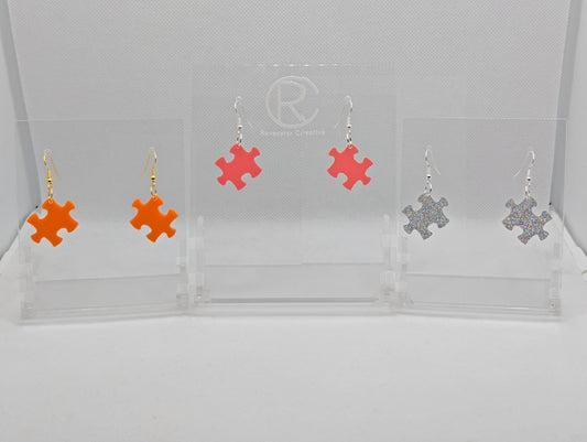 Shows three pairs of acrylic earrings with ear wire hook fixings. They are Jigsaw Game Piece Shaped. The colours are Orange, Holographic Glitter and Sherbet Pink. They are against a white background on a clear stand.