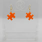 Shows a pair of acrylic earrings with gold ear wire hook fixings. They are Jigsaw Game Piece Shaped. The colour is Orange. They are against a white background on a clear stand with the Ravenstor Creative logo on it.