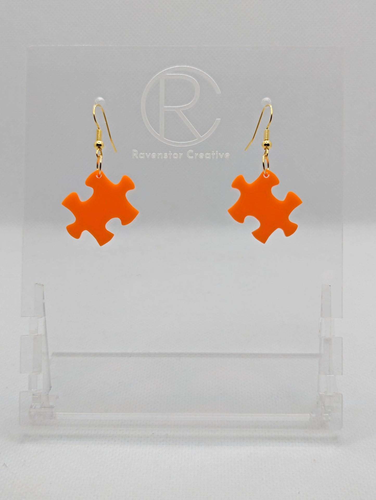 Shows a pair of acrylic earrings with gold ear wire hook fixings. They are Jigsaw Game Piece Shaped. The colour is Orange. They are against a white background on a clear stand with the Ravenstor Creative logo on it.