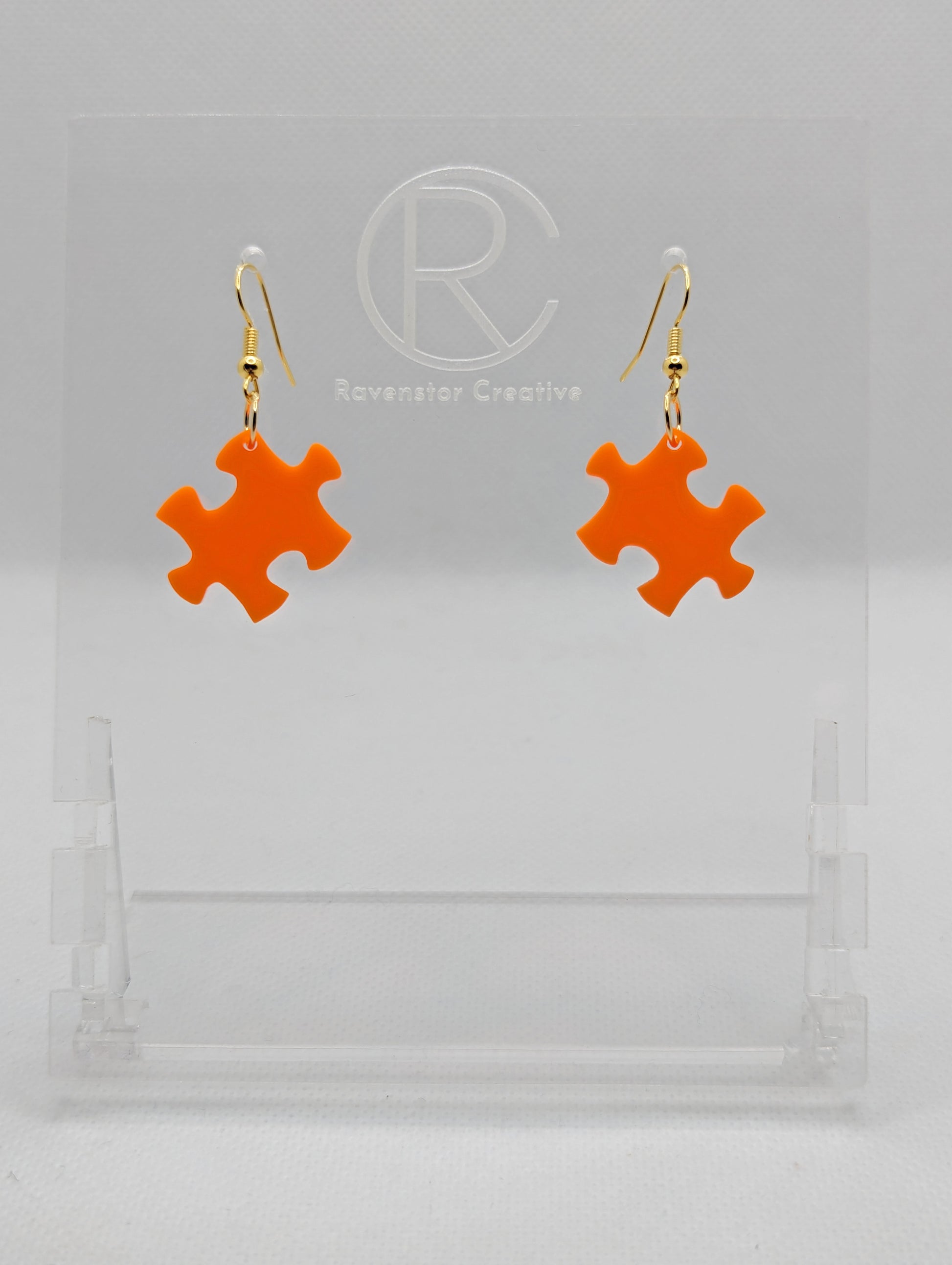 Shows a pair of acrylic earrings with gold ear wire hook fixings. They are Jigsaw Game Piece Shaped. The colour is Orange. They are against a white background on a clear stand with the Ravenstor Creative logo on it.