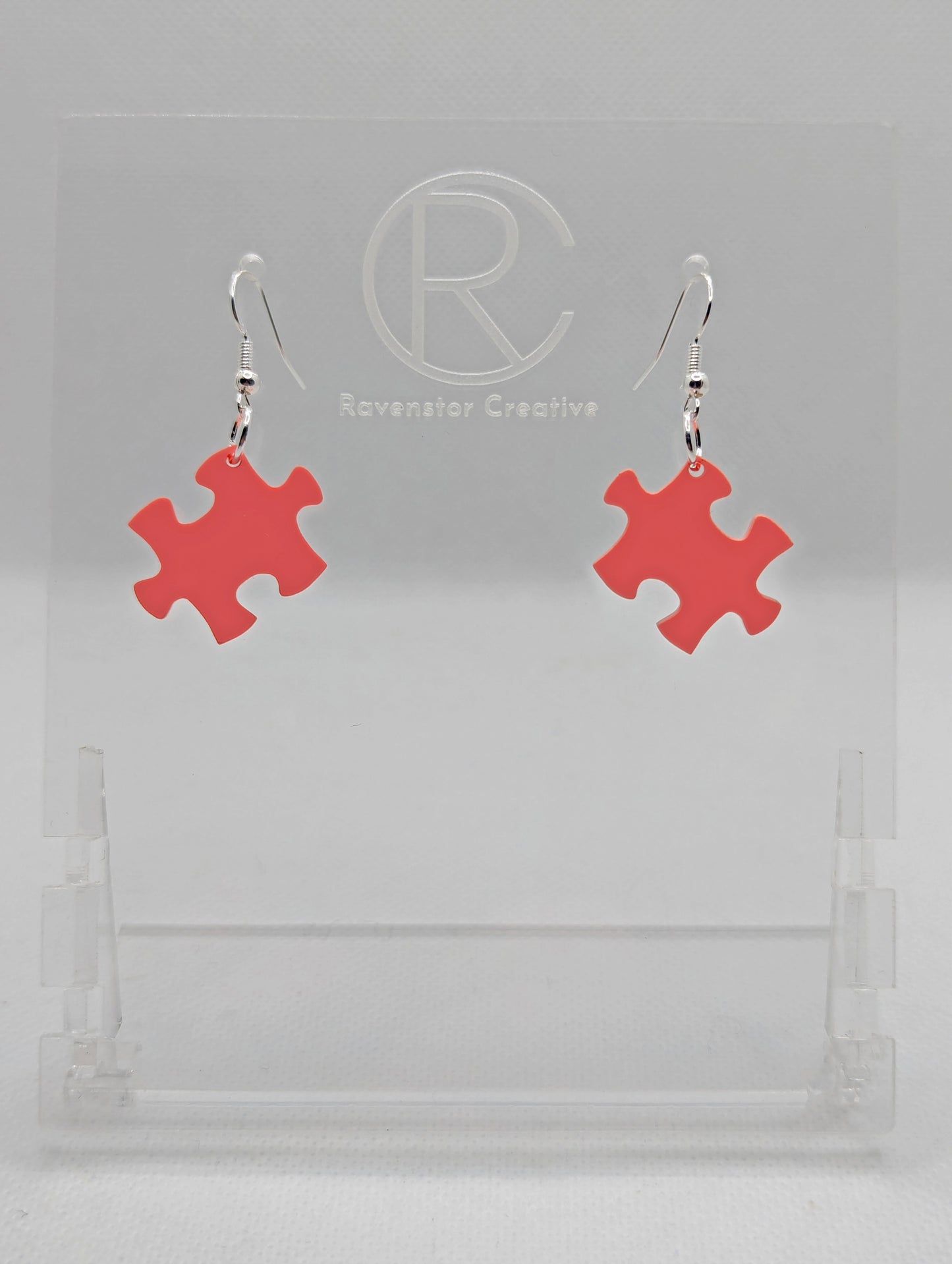 Shows a pair of acrylic earrings with silver ear wire hook fixings. They are Jigsaw Game Piece Shaped. The colour is Sherbet Pink. They are against a white background on a clear stand with the Ravenstor Creative logo on it.