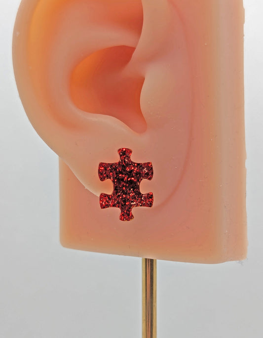 Shows a pair of small studded acrylic earrings that are a Jigsaw Game Piece Shape. They are Red Glitter in colour. They are on an ear to show the size.