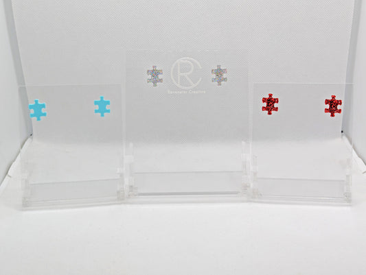 Shows three pairs of small studded acrylic earrings that are a Jigsaw Game Piece Shape. The colours are Spearmint, Holographic Glitter and Red Glitter. They are against a white background on a clear stand with the Ravenstor Creative logo on it.