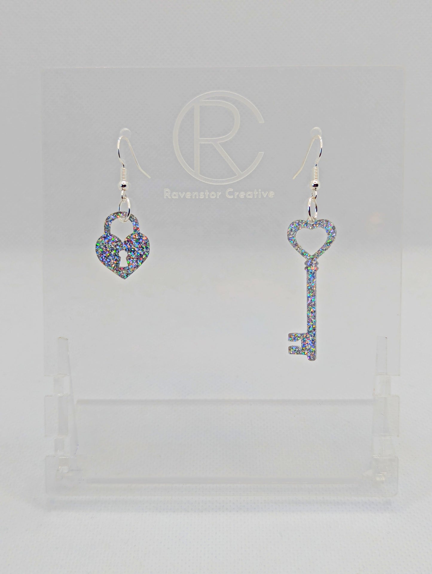 A set of two part drop earrings with Silver ear wire hook fixings. One earring is a Lock and the other is a Key, they are heart themed. The colour for both is Holographic Glitter. They are on a clear stand against a white background.