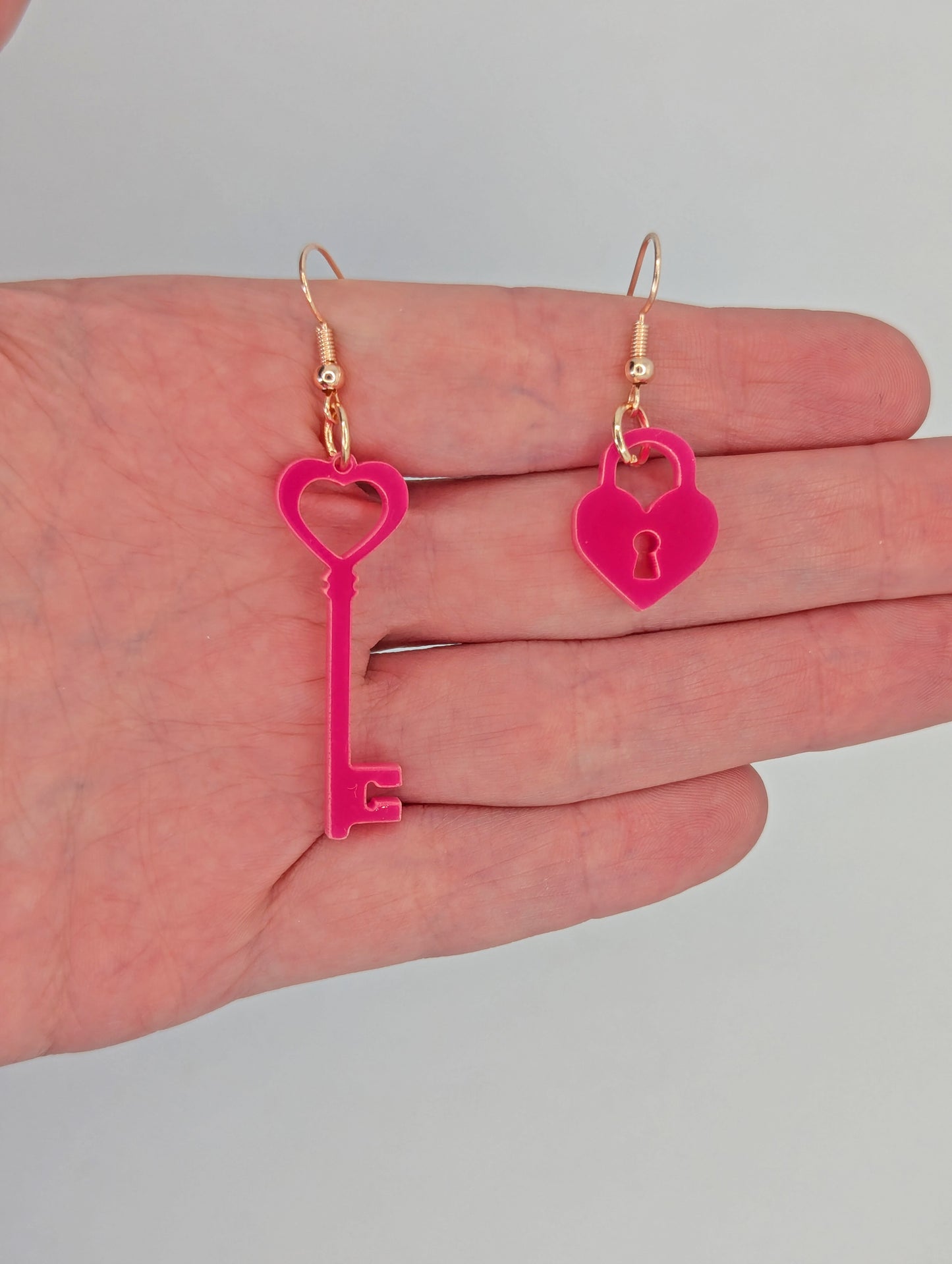 A set of two part drop earrings with rose gold ear wire hook fixings. One earring is a Lock and the other is a Key. They are against a hand to show the sizes. The key is two finger widths long and one wide. The Lock is one finger width long and wide.