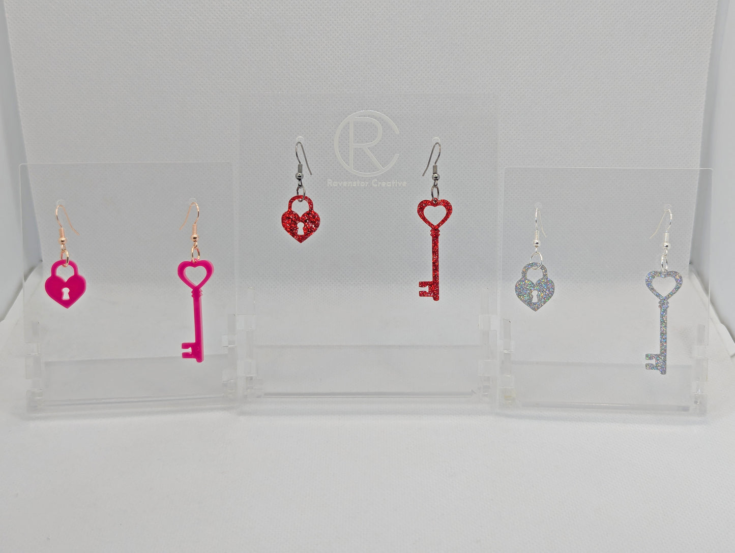 Three pairs of Two part drop earrings with ear wire hook fixings. Each set is a Lock and a Key. The colours are Hot Pink, Red Glitter and Holographic Glitter. They are on clear stands with the Ravenstor Creative logo against a white background.