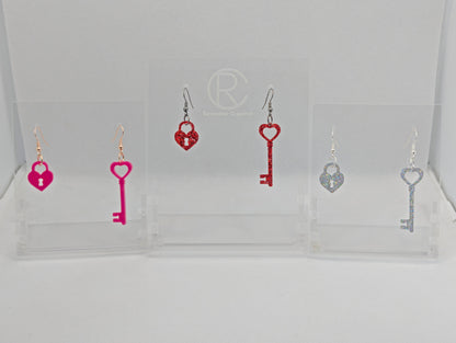 Three pairs of Two part drop earrings with ear wire hook fixings. Each set is a Lock and a Key. The colours are Hot Pink, Red Glitter and Holographic Glitter. They are on clear stands with the Ravenstor Creative logo against a white background.