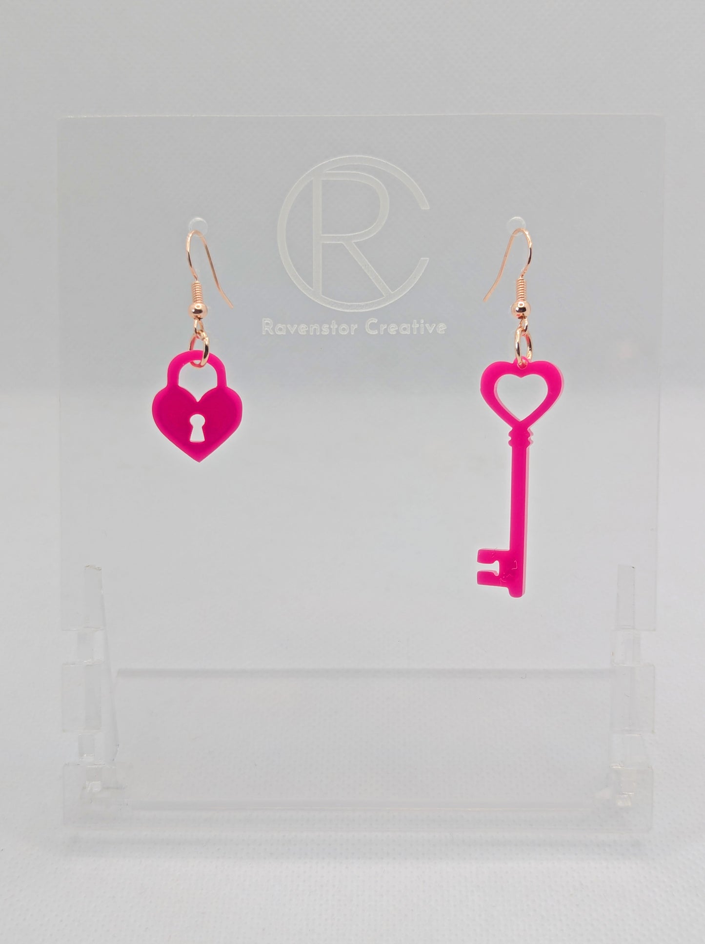 A set of two part drop earrings with Rose Gold ear wire hook fixings. One earring is a Lock and the other is a Key, they are heart themed. The colour for both is Hot Pink. They are on a clear stand against a white background.