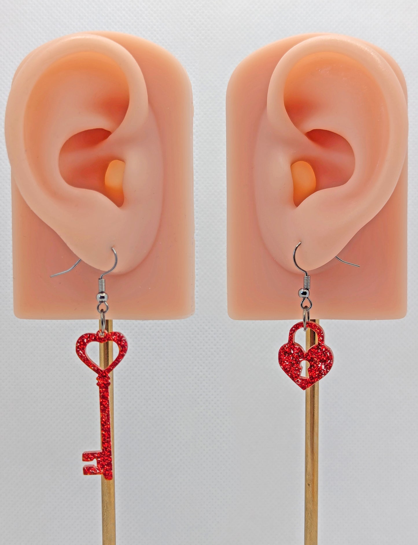 A set of two part drop earrings with Black ear wire hook fixings. One earring is a Lock and the other is a Key, they are heart themed. The colour for both is Red Glitter. They are on ears to show the size of each one.