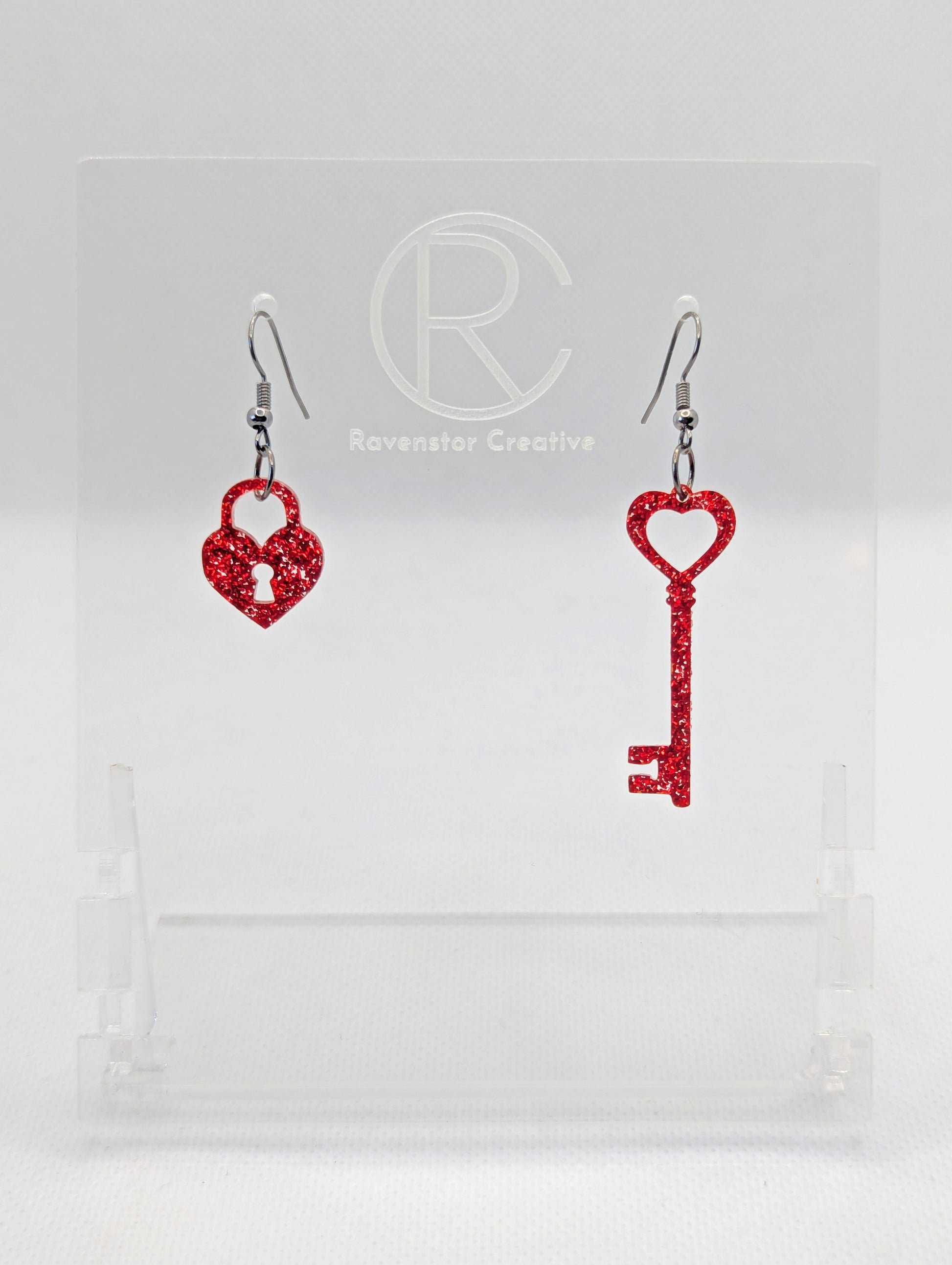 A set of two part drop earrings with Black ear wire hook fixings. One earring is a Lock and the other is a Key, they are heart themed. The colour for both is Red Glitter. They are on a clear stand against a white background.