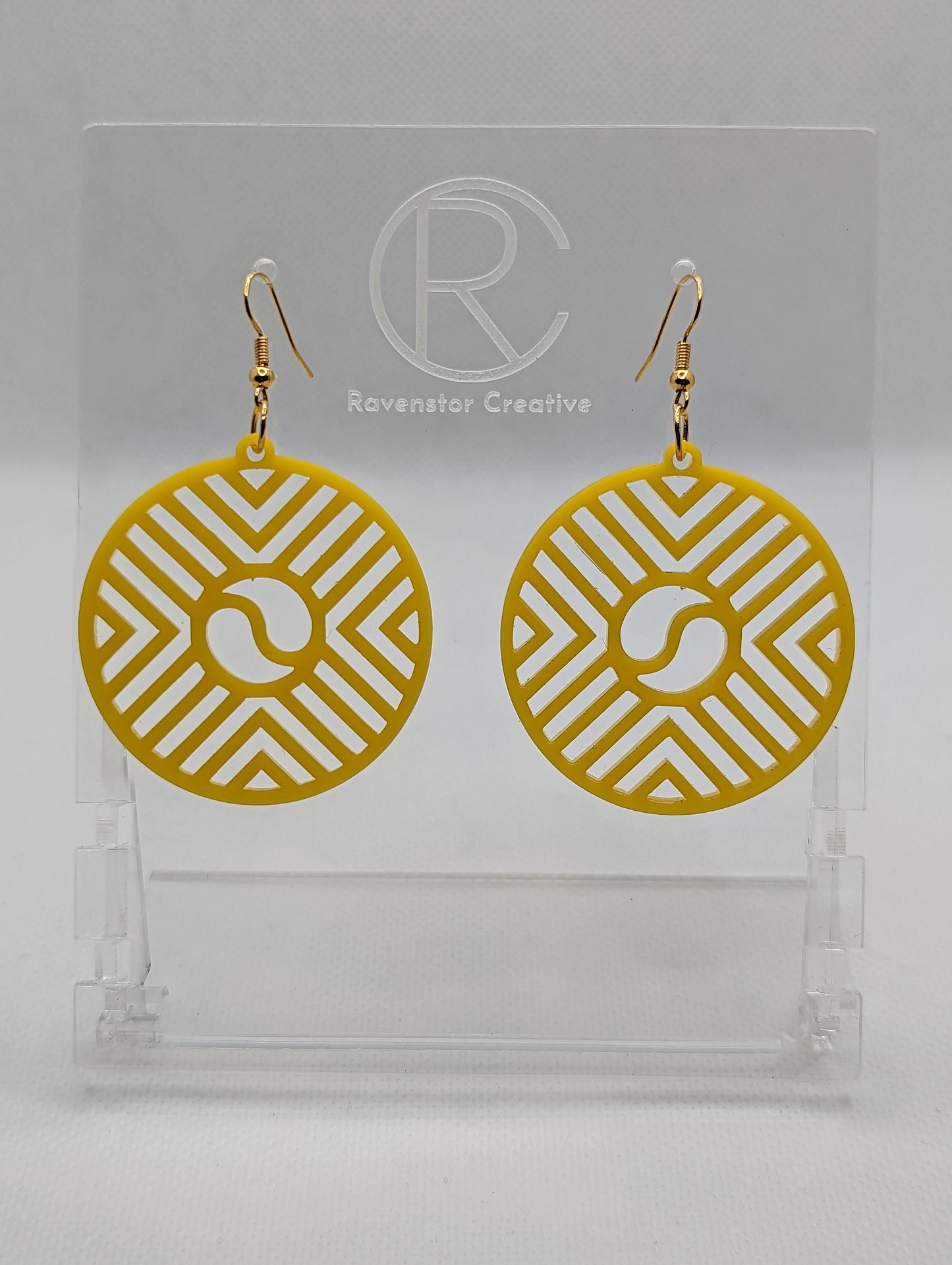 Shows a pair of Circle earrings that have gold ear wire fixings. They are Geometric Chevron designs with a yin and yang in the centre. They are made from yellow Acrylic. They are against a white background on see through stands.