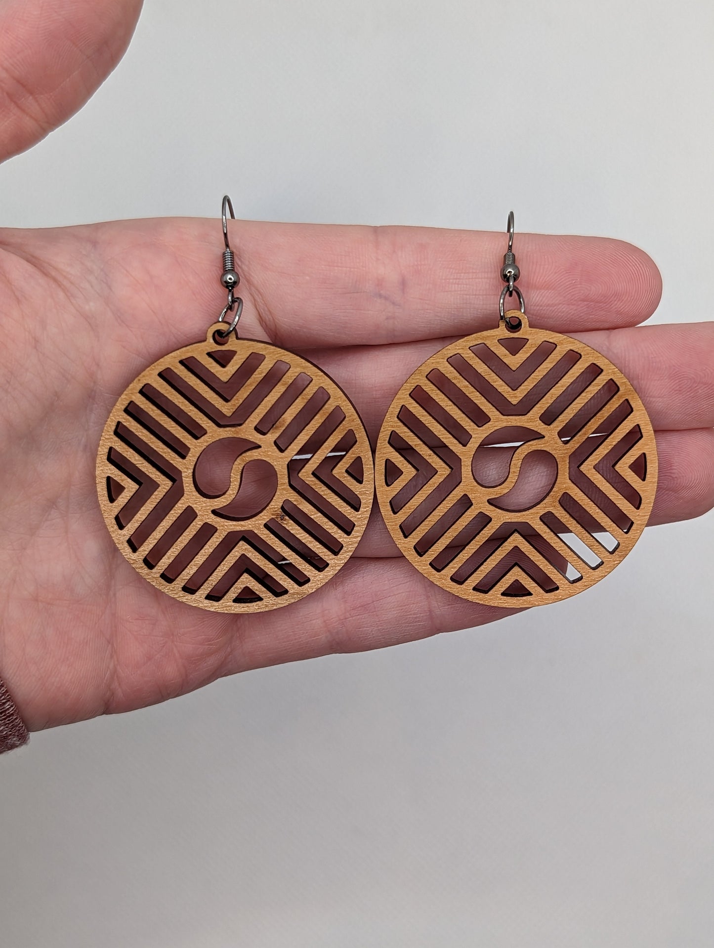 Image of a pair of Circle earrings with a Geometric Chevron design on a hand showing the size against a white background, they are three finger widths in length and three wide. They are Cherry Wood and have black fixings.