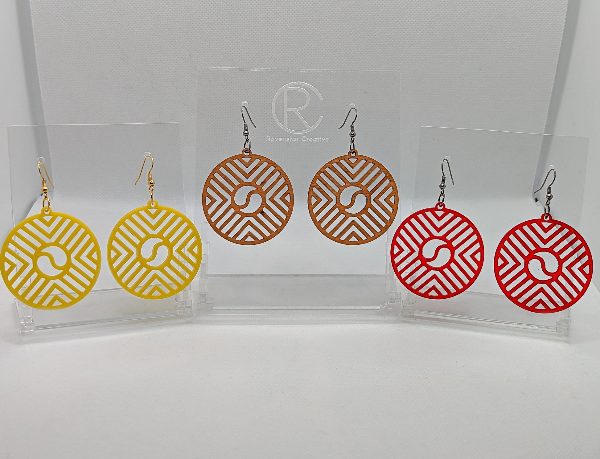 Shows three pairs of Circle earrings that have ear wire fixings. They are Geometric Chevron designs with a yin and yang in the centre. The colours are Yellow, Cherry Wood and Red. They are against a white background on see through stands.